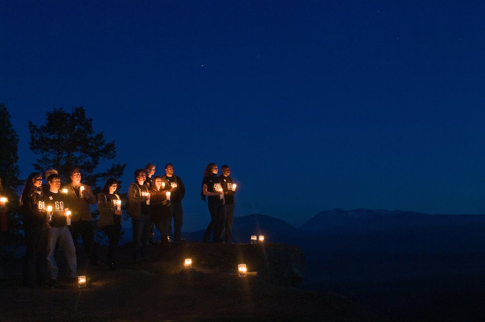  A group of men and women gathering on a clifftop on Vancouver Island against silhouetted trees and a twilight sky looking toward the sky and forested scenery, Earth Hour 2010, Canada. 